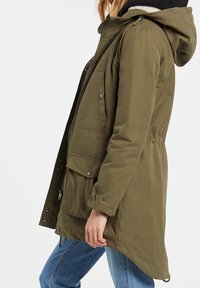 Parka vert olive avec capuche, dotée de boutons-pression, poches avant et taille cintrée. Tissu texturé avec une finition lisse.