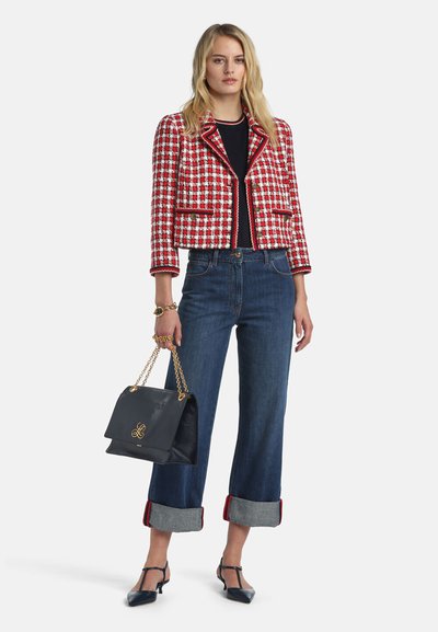 Žena u crveno-bijeloj kockastoj skraćenoj jakni, crnoj majici, plavim trapericama s podvrnutim nogavicama, crnim štiklama i drži crnu torbu s ručkom od zlatnog lanca.