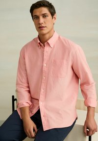 Młody mężczyzna ubrany w różową, zapinaną na guziki koszulę z długim rękawem i ciemne spodnie, siedzący na jasnym krześle na neutralnym tle.