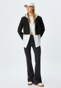 Veste noire coupée avec capuche grise, chemise blanche et pantalons noirs évasés avec rayures sur les côtés. Une casquette blanche et des baskets beiges complètent le look.
