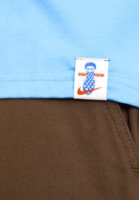 Light blue T-shirt with a white label featuring a cartoon character and "SOLE FOOD." Paired with brown textured pants.