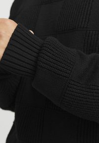 Melnā adīta džempera dizains iekļauj teksturētu šaha laukuma rakstu un ribotas piedurknes. Materiāls izskatās mīksts ar jauku apdari.