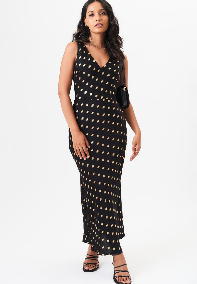 Black sleeveless dress with a V-neck, featuring gold oval polka dots, fitted silhouette, and a flowing hem. Styled with strappy black sandals.