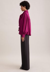 Blusa fucsia con maniche oversize e scollo arricciato, abbinata a pantaloni neri su misura, caratterizzati da un tessuto liscio e un design elegante.