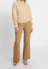 Maglione lavorato a maglia beige con texture a coste, abbinato a pantaloni a zampa d'elefante marroni a scacchi con righe gialle. Sandali morbidi color beige completano il look.