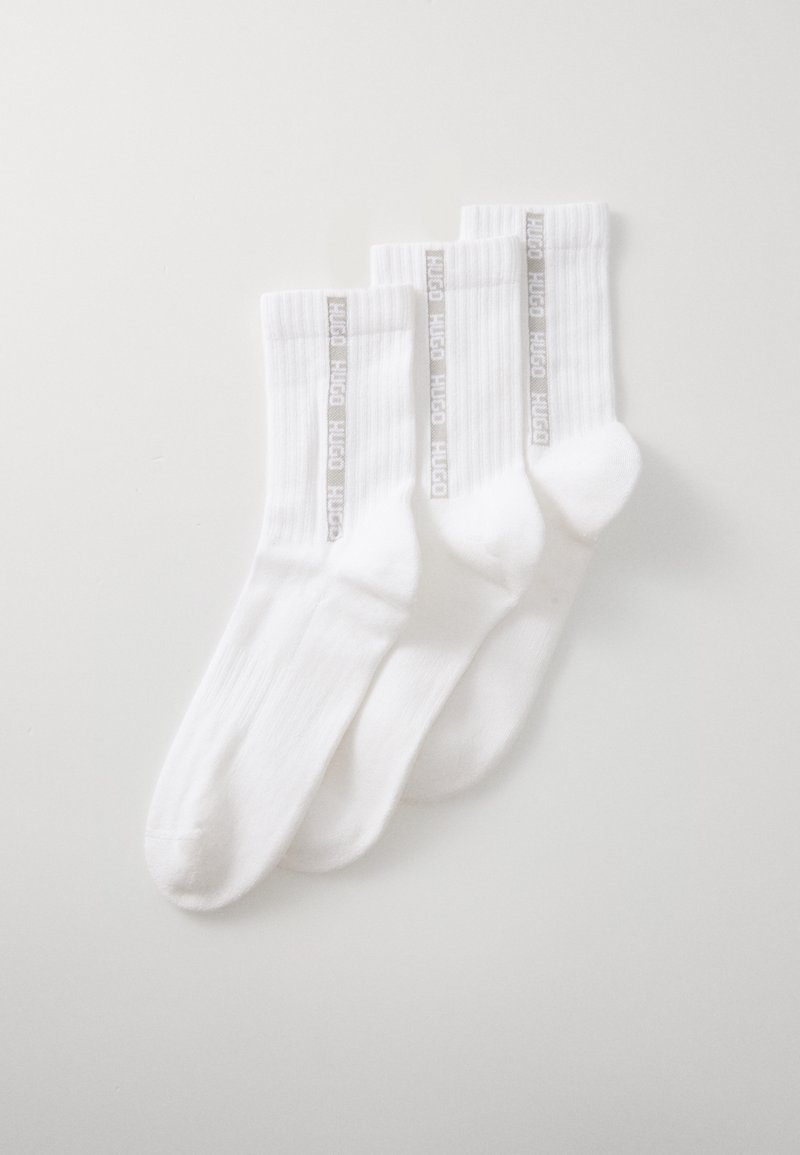Trois paires de chaussettes en coton blanc avec une texture côtelée et une bande de marque grise portant l'inscription "HUGO" verticalement sur chaque chaussette.