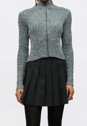 Person wearing a gray ribbed zip-up sweater, black pleated skirt, and black tights against a plain light background.