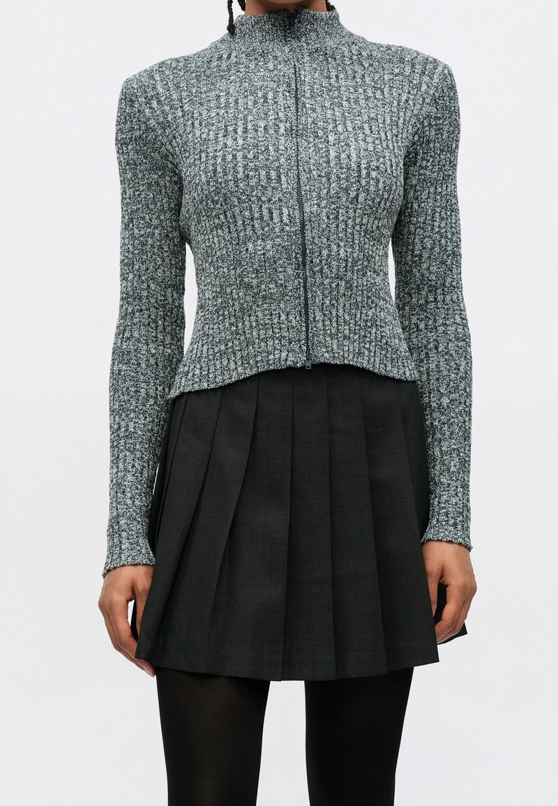 Personne portant un pull à fermeture éclair gris côtelé, une jupe plissée noire et des collants noirs sur un fond clair uni.
