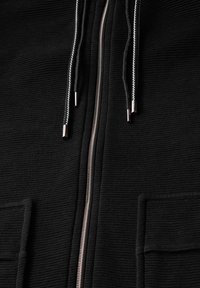 Schwarze Kapuzenjacke mit strukturiertem Stoff, ausgestattet mit einem silbernen Reißverschluss und Kordeln sowie zwei frontalen Taschen. Nahaufnahme des Kleidungsstücks.