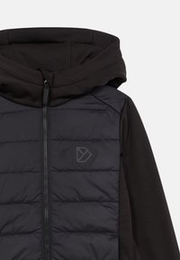 Melna pūkainā veste ar gludu tekstūru, kas papildināta ar pelēku logotipu, ar rāvējslēdzēja aizdari un piestiprinātu melnu kapuci no kontrastējoša materiāla.