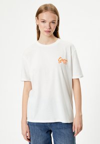 Hvid bomulds T-shirt med rund halsudskæring, korte ærmer og et orange grafisk motiv "California 98" på venstre bryst. Bæres med blå jeans.