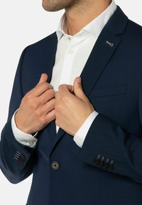 Man die een donkerblauwe colbert over een wit overhemd past, met details van knopen en kraag, gecropt bij de nek en taille.