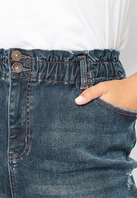 Jeans i denim med elastisk midja, knappdetalj, sidoficka, mörk tvätt och synliga stickningsaccents. Närbild av tygstruktur.