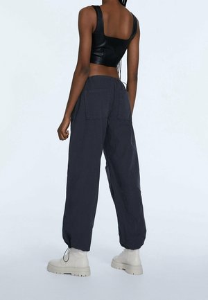 Donna che indossa un top corto in pelle nera, pantaloni grigi scuri larghi con tasche posteriori e stivali bianchi chunky, che sta in piedi contro uno sfondo neutro.