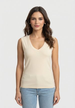 Femme aux cheveux bruns ondulés, portant des boucles d'oreilles en perles, un haut beige sans manches à col en V, et un jean bleu clair, debout avec les bras détendus le long du corps.