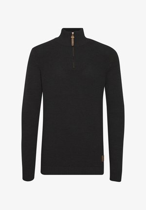 Melnā, adīta džemperis ar augstu apkakli, pusi rāvējslēdzēja aizdari un ribotu tekstūru. Tam ir mazs, kvadrātveida zīmola birka apakšējā malā.