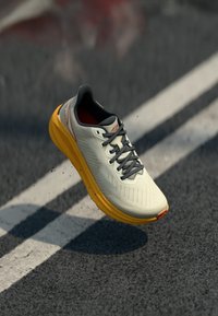 Leichter Laufschuh mit einem cremefarbenen Obermaterial, strukturiertem Design, grauen Schnürsenkeln und einer hellgelben Sohle. Hervorstechendes rotes Detail an der Ferse.