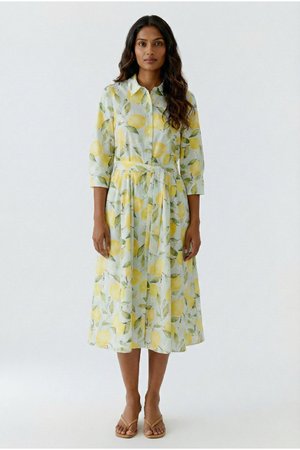Femme portant une robe midi à manches longues avec un imprimé citron jaune et feuilles vertes, debout devant un fond blanc uni.