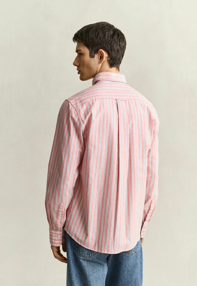 Homme portant une chemise à manches longues rayée rose et blanche boutonnée et un jean bleu, de dos face à un fond uni clair.