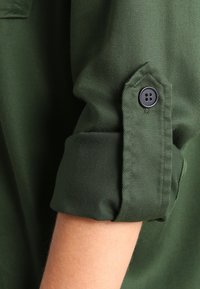 Camicia verde con maniche arrotolate, dotata di dettagli con bottoni. Tessuto liscio con una finitura leggermente strutturata.