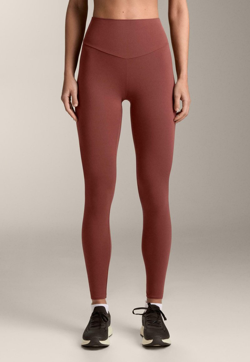 Donna che indossa leggings color ruggine a vita alta e scarpe sportive nere con calzini bianchi alla caviglia, in piedi davanti a uno sfondo neutro.