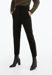 Černé kalhoty se slim fit střihem, detailem opasku a nýty po straně. Vyrobené z hladké látky, doplněné o špičaté černé boty na podpatku.