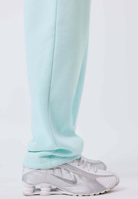 Persona che indossa pantaloni azzurri chiari e scarpe da ginnastica bianche Nike Shox, in piedi su uno sfondo bianco.