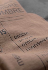 Vaaleanruskea kangas, jossa on mustalla painetut tekstit, toistuvat sanat "ombre" ja "menswear" yksinkertaisella, lihavoidulla fontilla. Pehmeä rakenne.