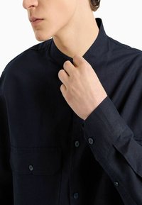 Camicia blu scuro con colletto alla coreana, due tasche anteriori e chiusure con bottoni. Realizzata in tessuto lucido con finitura testurizzata e maniche lunghe.
