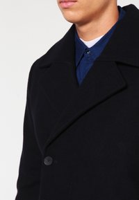 Schwarzer Wollmantel mit Reverskragen, Einzelknopfverschluss und breiten Schultern. Wird über einem blauen Hemd mit Kragen getragen. Glatte Textur.