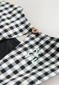 Mustvalge ruuduline kangas, millel on nupuga sulgemine ja tikitud helesinise tekstiga detail. Sile tekstuur ja kohandatud kuju.