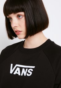 Sudadera negra con el logo blanco de "VANS", cuello de canalé y mangas raglán. La tela parece suave y cómoda.