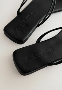 Sandales plates noires avec des fines lanières, présentant une texture matte lisse et un logo de marque en relief. La semelle est plate et arrondie au bout.