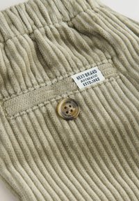 Jasnozielony materiał kordowy z pionowymi żeberkami, z brązowym guzikiem i metką z napisem "HEAT BRAND AUTHENTIC ESTD. 1982."