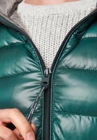 Grøn pufferjakke med høj krave, lynlåslukning og elastisk snøre. Båret over en lys grå tekstureret striktrøje.