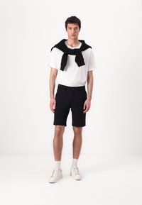 Black shorts with a fitted design, paired with a white t-shirt and a black sweater tied around the shoulders, worn with white sneakers.