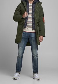 Olive green parka with a detachable hood and navy lining, layered over a striped sweater. Worn with distressed jeans and white sneakers.