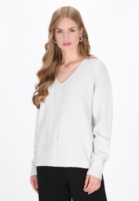 Jasnoszara sweter w serek wykonany z miękkiej dzianiny, z teksturowanym wzorem i ściągaczami przy mankietach, zestawiony z czarnymi spodniami.