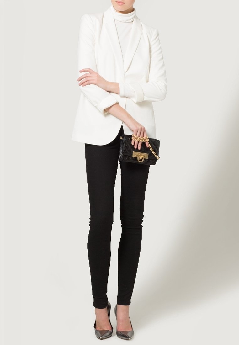 Osoba w białym żakiecie i golfie, czarnych spodniach, metalicznych czubkach na obcasie, trzymająca czarną torebkę z złotym łańcuchem.