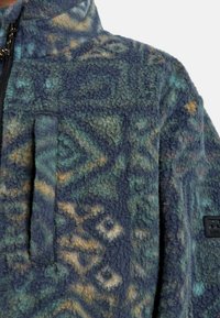 Fleecejacke mit einem gemusterten Design in Dunkelblau, Grün und erdigen Tönen, mit einem Reißverschluss und strukturierter Oberfläche.