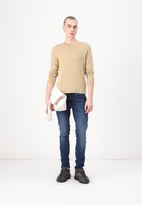 Beige strikkegenser med ribbede mansjetter, blå slim-fit jeans og svarte teksturerte joggesko. Holder en kremfarget veske.