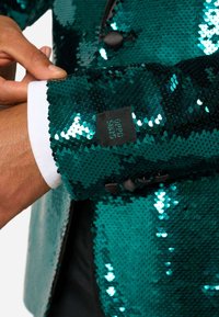 Błyszcząca marynarka w kolorze niebiesko-zielonym z cekinami, z kontrastującymi czarnymi guzikami i białą mankietą wystającą spod rękawa. Metka widoczna na mankiecie.