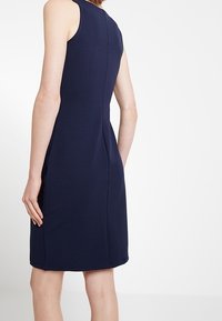 Granatowa sukienka bez rękawów o dopasowanym kroju, z detalami na szwie tylnym i gładką strukturą materiału. Długość do połowy uda.