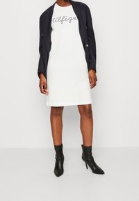 White dress with printed text, paired with a navy blazer and black heeled ankle boots. Smooth fabric, simple silhouette, and tailored fit.