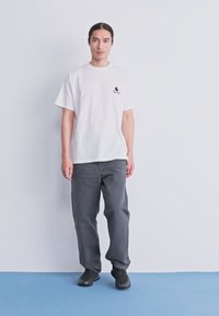 Giovane uomo con lunghi capelli scuri, indossa una maglietta bianca con grafiche, pantaloni grigio scuro e scarpe nere, in piedi su un pavimento blu contro un muro bianco.