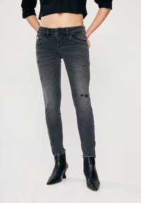 Černé zkrácené džíny s odřenými detaily a slim fit. Vyrobené z denimové tkaniny, mají knoflíkový zip a pět kapes. Nohavice jsou kombinovány s černými špičatými botami.