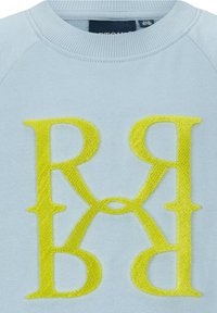 Jasnoniebieska bluza z prążkowanym dekoltem, z dużymi haftowanymi żółtymi literami "R" i "B" w symetrycznym wzorze.