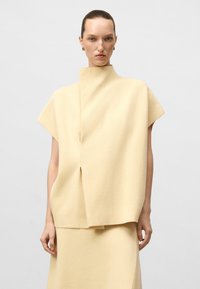 Femme avec les cheveux tirés en arrière portant un haut ample à col montant et à manches courtes de couleur jaune pâle, et une jupe assortie, sur un fond uni.