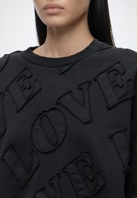 Gros plan d'une personne portant un sweat-shirt noir avec de grandes lettres texturées formant le mot « LOVE » à l'avant.
