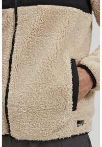 Beige nepbont jasje met zwarte accenten, voorzien van een rits, zijzakken en een logo patch aan de onderkant. Zachte, gestructureerde oppervlakte.